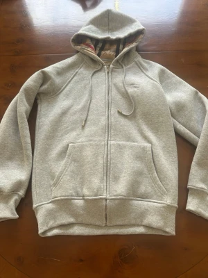 Grå Burberry zip hoodie  - Säljer en stilren grå hoodie med dragkedja och stor ficka framtill. Hoodien har justerbar huva med snören och ribbade muddar vid ärmar och nederkant. Insidan av huvan har ett rutigt tyg som detalj. Perfekt för en avslappnad och bekväm look.