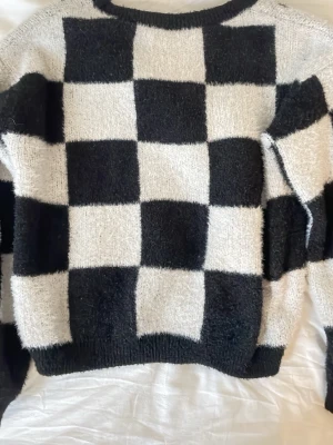 Stickad tröja med rutigt mönster - Säljer en stickad tröja med svart och vitt rutigt mönster. Tröjan har en mjuk, fluffig känsla och raka ärmar. Perfekt för dig som gillar en trendig och avslappnad stil. Materialet känns mysigt och passar bra till höst och vinter.