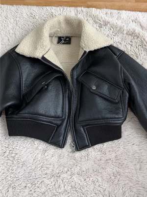 Svart skinnjacka med teddykrage - Snygg svart skinnjacka med vit teddykrage och foder. Jackan har två stora fickor med lock och tryckknapp framtill, samt ribbade muddar i midja och ärmslut. Kortare modell som ger en cool och trendig look.