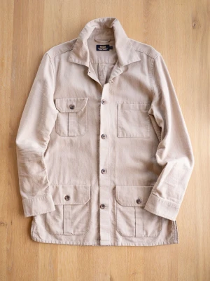Beige Wacay Field Jacket, size S - Stilren beige overshirt i kraftigt bomullstyg med fyra stora fickor framtill och knäppning med bruna knappar. Klassisk krage och raka ärmar ger en avslappnad look. Perfekt att bära över en t-shirt eller hoodie för en trendig lager-på-lager stil. Liten fläck på ryggen. 