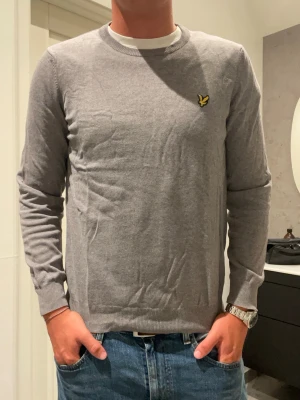 Grå tröja från Lyle & Scott - Snygg grå tröja från Lyle & Scott med rund halsringning och den klassiska gula loggan på bröstet. Tröjan har långa ärmar och ribbade muddar vid ärmslut och nederkant. Perfekt till jeans eller chinos.