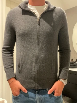 Grå ziptröja från Polo Ralph Lauren - Mjuk och stilren grå ziptröja från Polo Ralph Lauren. Tröjan har hög krage, dragkedja framtill och två fickor. Liten broderad logga på bröstet. Perfekt till jeans eller över en t-shirt för en avslappnad look.