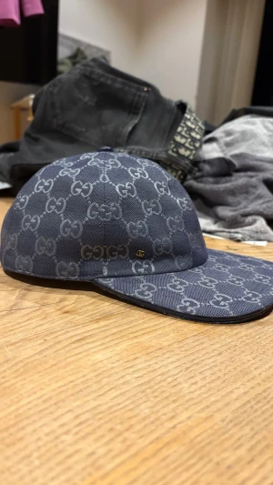 Blå gucci keps - Snygg blå keps från Gucci med det klassiska GG-monogrammönstret över hela kepsen. Tillverkad i slitstarkt tyg med böjd skärm och justerbar baksida. Insidan är ljus och har etikett med Gucci-logga. Perfekt för dig som gillar exklusiva accessoarer.