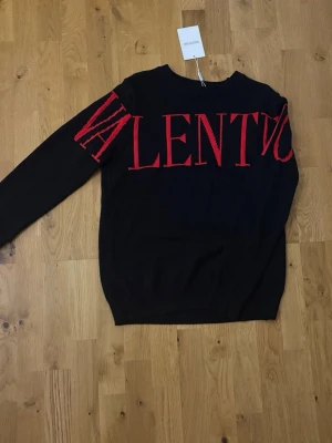 Valentino tröja - Svart stickad tröja från Valentino med stora röda bokstäver över bröstet. Tröjan har rund halsringning och ribbstickade muddar. Tillverkad i Italien och har en modern, oversized look. Perfekt för dig som vill sticka ut med en exklusiv design.