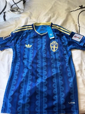 Blå Sverige fotbollströja Adidas - Säljer en blå fotbollströja från Adidas med Sveriges landslagsemblem på bröstet. Tröjan har gula detaljer, klassiska Adidas-ränder på axlarna och ett unikt mönster över hela tyget. Tillverkad i lätt och ventilerande material med Climalite-teknologi. Passar även s