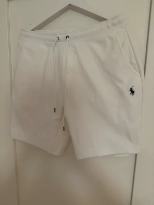 Vita shorts från Polo Ralph Lauren - Säljer ett par fräscha vita shorts från Polo Ralph Lauren med snörning i midjan och fickor på sidorna. Klassisk logga broderad i mörkblått på ena benet. Sköna och stilrena, perfekta för sommaren.