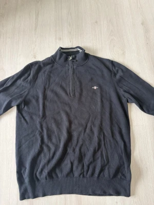 Mörkblå stickad tröja från GANT - Mörkblå stickad tröja från GANT med halv dragkedja och liten broderad logga på bröstet. Tröjan har ribbade muddar och krage med vit randdetalj. Perfekt för en stilren och avslappnad look.