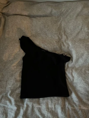 Svart oneshoulder topp Stradivarius - Snygg svart oneshoulder topp från Stradivarius. Toppen har en asymmetrisk design med ena axeln bar och ett smalt axelband på andra sidan. 🖤