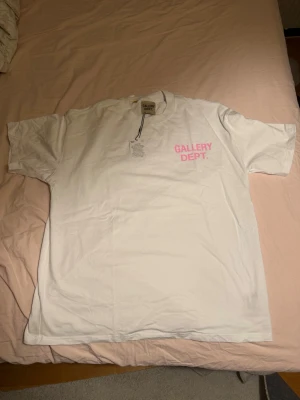 Vit Gallery Dept. t-shirt med rosa tryck - Säljer en vit t-shirt från Gallery Dept. med rosa texttryck både fram och bak. T-shirten har klassisk passform, rund hals och korta ärmar. Materialet är mjuk bomull som känns skön mot huden. Perfekt för dig som gillar streetwear och vill sticka ut.