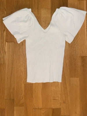 Vit ribbad topp med puffärm - Säljer en vit ribbad topp med djup v-ringning och stora puffärmar. Toppen har en figurnära passform och är tillverkad i ett stretchigt material som känns mjukt mot huden. Perfekt för dig som vill ha en trendig och stilren look.