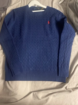 Blå kabelstickad tröja Polo Ralph Lauren - Snygg blå kabelstickad tröja från Polo Ralph Lauren med klassisk röd logga broderad på bröstet. Tröjan har rund halsringning, långa ärmar och ribbade muddar. Perfekt för dig som vill ha en stilren och tidlös look. Står L men sitter som M