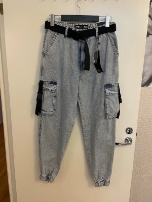 Grå cargobyxor med bälte FB SISTER, stl M nya  - Snygga grå cargobyxor från FB SISTER i tvättad denim-look. Byxorna har stora sidofickor med lock, elastiska benslut och ett svart justerbart bälte med plastspänne i midjan. Perfekta för en avslappnad streetwear-stil.