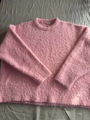 Ljusrosa fluffig stickad tröja - Supermjuk och fluffig stickad tröja i ljusrosa färg. Tröjan har rund halsringning, långa ärmar och en avslappnad passform. Perfekt för att hålla sig varm och mysig under kyliga dagar.