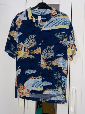 Blå hawaiiskjorta med palmmotiv - Säljer en blå kortärmad hawaiiskjorta med färgglatt palmmotiv och vågor. Skjortan har klassisk krage och knappar framtill. Perfekt för sommaren och ger riktigt chill semesterkänsla.
