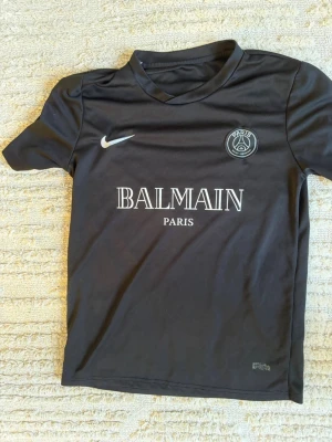 Svart PSG x Balmain t-shirt - Snygg svart t-shirt med PSG och Balmain Paris-logga på bröstet. Tröjan har rund hals, korta ärmar och är tillverkad i ett mjukt, sportigt material. Perfekt för dig som gillar fotboll och streetwear.