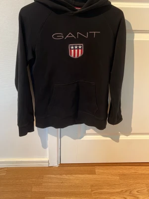 Svart hoodie från GANT med logga - Säljer en svart hoodie från GANT med stor huva och känguruficka. På bröstet finns GANT-loggan och ett broderat märke i rött, vitt och blått med stjärnor och ränder. Hoodien har långa ärmar och är gjord i mjukt material, perfekt för en chill och stilren look. Storlek 13-14 år 158/164.