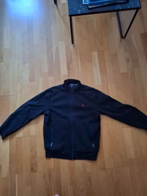 Svart bomberjacka från Polo Ralph Lauren - Klassisk svart bomberjacka från Polo Ralph Lauren med dragkedja framtill och två fickor med dragkedja. Jackan har en liten röd logga broderad på bröstet och ribbade muddar vid ärmslut och nederkant. Perfekt för en stilren och avslappnad look.