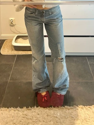 Ljusblå bootcut jeans med slitning - Säljer ett par ljusblå bootcut/baggy jeans med slitning och hål på ena benet. Jeansen har klassisk femficksmodell, låg midja och är tillverkade i denim. Perfekta för en avslappnad och trendig look.