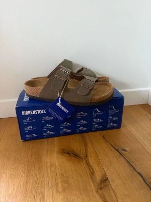 Bruna Birkenstock sandaler med spännen - Säljer ett par klassiska Birkenstock sandaler i oliv grön färg  med två breda remmar och justerbara metallspännen. Fotbädden är i mjukt material och sulan är i kork för extra komfort. Perfekta för en avslappnad och trendig stil.