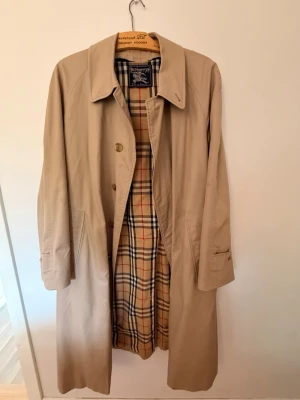 Beige trenchcoat från Burberrys - Klassisk beige trenchcoat från Burberrys med det ikoniska rutiga fodret i beige, svart, rött och vitt. Lång modell med krage och knäppning framtill. Tillverkad i England och har en tidlös, stilren look som passar perfekt till lager-på-lager.