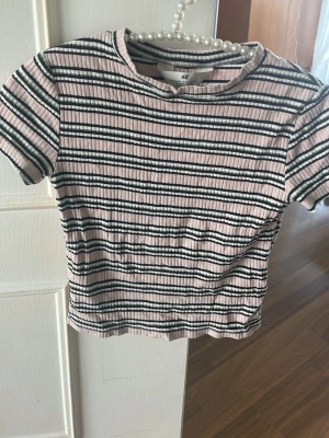 Ribbad randig t-shirt från H&M - Säljer en ribbad t-shirt från H&M med svart, vit och ljusrosa randigt mönster. T-shirten har korta ärmar och en klassisk rund halsringning. Perfekt för dig som gillar en enkel men trendig stil.