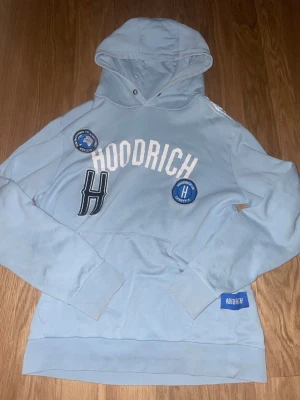 Ljusblå hoodie från Hoodrich - Snygg ljusblå hoodie från Hoodrich med vita och blå broderade patches och tryck framtill. Hoodien har huva, magficka och ribbade muddar. Perfekt för en chill och streetinspirerad look.
