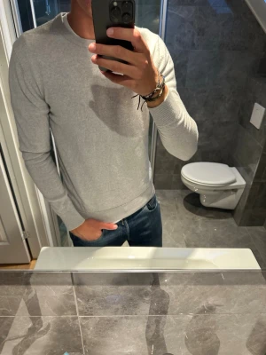 Ljusgrå tröja i bomull John Henric - En stilren ljusgrå tröja med rund hals från John Henric . Tröjan är tunnstickad och har ribbade muddar vid ärmslut och nederkant. Väldigt användbar då den passar till det mesta. Tröjan är i stort sätt bra skick bortsett från en lite fläck på ärmen (se sista bild). Hör av er vid fler frågor:)