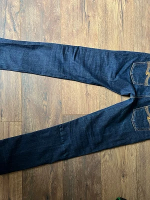 Mörkblå jeans från Nudie Jeans - Säljer ett par mörkblå jeans från Nudie Jeans i storlek 31/32. Klassisk rak passform med snygga kontrastsömmar och detaljerade bakfickor. Tillverkade i slitstarkt denimtyg som ger en tidlös look.