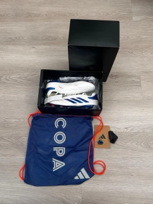 Adidas Copa vita och blå fotbollsskor - Säljer ett par Adidas Copa fotbollsskor i vitt med blå metallic detaljer och röda logotyper. Skorna har klassisk snörning, låg profil och är tillverkade i syntetmaterial och läder. Medföljer blå skopåse med Copa-tryck och extra tillbehör.