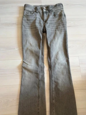 Grå bootcut jeans med dubbla knappar - Snygga grå bootcut jeans med dubbla silverknappar i midjan och klassiska fem fickor. Jeansen har en lätt tvättad look och utsvängda ben. Perfekta för dig som gillar en avslappnad men trendig stil.