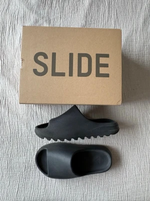 Svarta Adidas Yeezy Slide sandaler - Säljer ett par Adidas Yeezy Slide i mycket bra skick. De köptes för tre år sedan och har inte använts så mycket. Jag har authentication och lådan ingår också. De är riktigt sköna att gå med hemma och passar även till mörkare outfits.