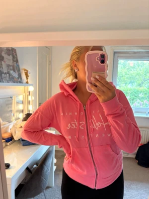 Rosa hoodie med dragkedja - Säljer en rosa hoodie med dragkedja och huva. Hoodien har tryck framtill med text och två fickor på magen. Perfekt för dig som gillar en avslappnad och sportig stil. Materialet ser mjukt och bekvämt ut, och modellen är klassisk med långa ärmar. Kom gärna med egna bud! 
