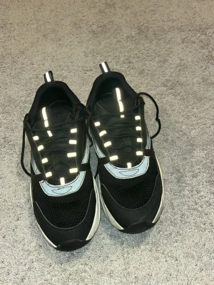 Svarta sneakers med reflexdetaljer - Säljer ett par svarta sneakers med vita och grå detaljer samt reflexdetaljer på snörningen. Skorna har meshpartier för extra ventilation och en vit sula som ger en sportig look. Perfekta för dig som gillar stilrena och funktionella sneakers.