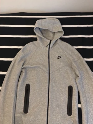 Grå Nike Tech Fleece hoodie - Säljer en grå Nike Tech Fleece hoodie med dragkedja framtill och två svarta dragkedjeförsedda fickor. Hoodien har en stilren design med Nike-logga på bröstet och är tillverkad i mjukt, stretchigt material. Perfekt för en sportig och avslappnad look.