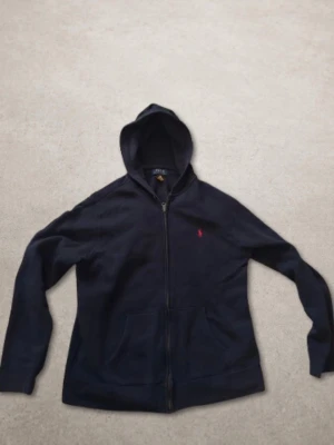 Mörkblå Ralph lauren hoodie med dragkedja - Mörkblå hoodie med dragkedja och huva. Klassisk design med fickor framtill och ribbade muddar vid ärmar och nederkant. Liten röd logga broderad på bröstet. Storleken är junior XL som motsvarar vuxen S.