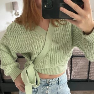 Ljusgrön omlottstickad tröja - Trendig ljusgrön stickad omlottopp med knytband i midjan och bred ribbad kant. Tröjan har lång ärm och en croppad passform som ger en modern look. Perfekt att styla med höga jeans för en avslappnad vibe.