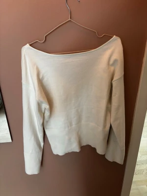 Beige stickad tröja med v-ringning - En enkel och stilren beige stickad tröja med djup v-ringning på ryggen och bred urringning fram. Tröjan har långa ärmar och en avslappnad passform, perfekt att matcha med jeans eller kjol. Materialet känns mjukt och lätt, vilket gör den skön att bära.