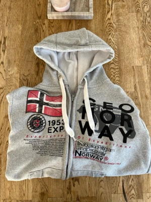 Grå hoodie från Geo Norway med tryck - Säljer en grå hoodie från Geo Norway med dragkedja, stor huva och vita snören. Framsidan har tryck med norsk flagga och text i svart och rött. Insidan är mjuk och luvan har ett vitt meshfoder. Finns några små tecken på användning