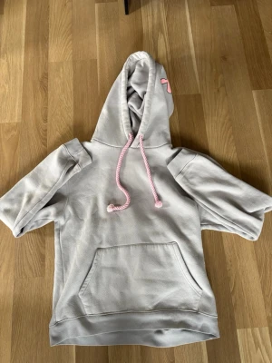 Ljusgrå hoodie med rosa detaljer - Säljer en ljusgrå hoodie med korta ärmar och magficka. Huvan har rosa snören och ett coolt rosa tryck på baksidan. Perfekt för dig som gillar streetwear och vill ha något unikt i garderoben.