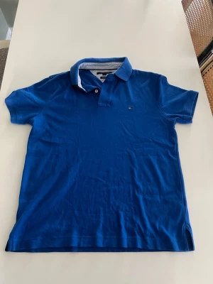 Blå pikétröja med krage - Säljer en klassisk blå pikétröja med krage och korta ärmar. Tröjan har knappar vid halsen och en liten logga på bröstet. Perfekt till jeans eller shorts för en avslappnad look.