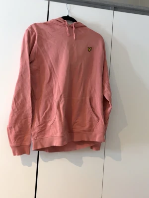 Rosa hoodie från Lyle & Scott - Säljer en rosa hoodie från Lyle & Scott med klassisk gul logga på bröstet. Hoodien har huva med dragsko och en stor magficka. Tillverkad i mjukt bomullsmaterial och har ribbade muddar vid ärmslut och nederkant. Perfekt för en chill och avslappnad stil.