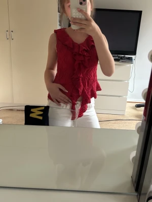 Röd volang topp med struktur - Säljer en röd ärmlös topp med volangdetaljer framtill och längs nederkanten. Toppen har en snygg struktur i tyget och är perfekt för dig som vill sticka ut lite extra. Passar till både jeans och kjol för en trendig look.