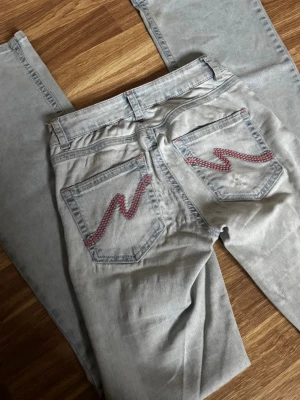 Ljusblå jeans med detaljer på fickorna  - Säljer ett par ljusblå jeans från Nelly med slitningar på knäna och rosa sicksack-detaljer på bakfickorna. Jeansen har klassisk femficksmodell, utsvängda ben och bälteshällor. Perfekta för en avslappnad och trendig look. Bra skick, inga defekter. Storlek 34. Pris går att diskutera💕💕