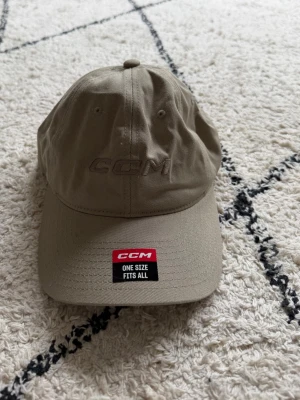 Beige keps från CCM - Nu beige keps från CCM med böjd skärm och broderad logga framtill. Justerbar passform med kardborreband baktill och ventilerande hål. Tillverkad i slitstark bomull. Helt ny och aldrig använd.