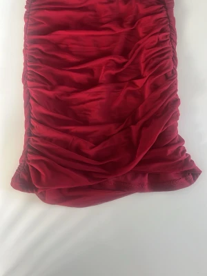 Röd kortkjol, funkar som top - Snygg röd kortkjol med omlottkänsla och rynkade detaljer längs sidorna. Kjolen har en kroppsnära passform och är tillverkad i ett mjukt, stretchigt material som ger en smickrande siluett. Perfekt för dig som vill sticka ut med färg.