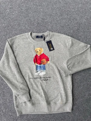 Grå sweatshirt Polo Bear Ralph Lauren - Säljer en grå sweatshirt från Ralph Lauren med den ikoniska Polo Bear-trycket framtill. Björnen har på sig en röd hoodie, blå jeans och vita sneakers samt håller i en basketboll. Tröjan har rund halsringning och ribbade muddar. Perfekt för en avslappnad och trendig look.
