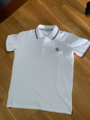 Vit Moncler pikétröja XL - Klassisk vit pikétröja från Moncler i storlek XL. Tröjan har korta ärmar, knappar framtill och Moncler-logga på bröstet. Kragen och ärmsluten har snygga ränder i rött och marinblått. Perfekt för en clean och stilren look.