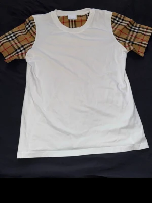 Vit t-shirt med rutiga ärmar - Snygg vit t-shirt med klassiska beige, svarta och röda rutiga ärmar och halsringning. T-shirten har rak passform och är tillverkad i mjukt bomullstyg. Perfekt för dig som vill sticka ut med en unik detalj på en annars enkel t-shirt.