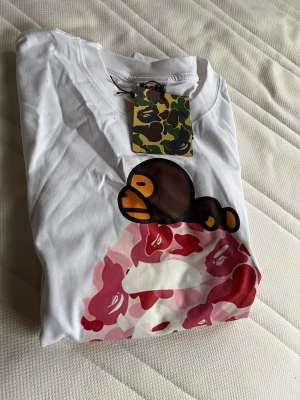 Vit BAPE t-shirt med apa och camo - Säljer en vit t-shirt från BAPE med ikoniskt tryck av en brun apa och rosa-röd camo-mönster på framsidan. T-shirten har korta ärmar och är i mjuk bomull. Perfekt för dig som gillar streetwear och vill sticka ut med en unik design.
