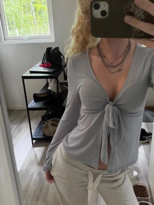 Ljusgrå långärmad topp med knyt - Trendig ljusgrå långärmad topp med djup v-ringning och knyt framtill. Tunt och mjukt material som sitter snyggt på kroppen. Perfekt för dig som gillar en avslappnad men ändå stilren look.